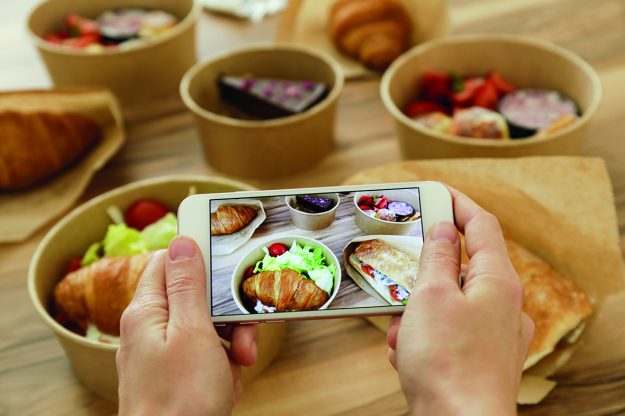 Female food blogger reviewing takeout food, taking video on phone. Italian sub sandwich, french croissant w/ salmon, chocolate cheesecake. Close up, top view, pov, copy space, wooden table background.