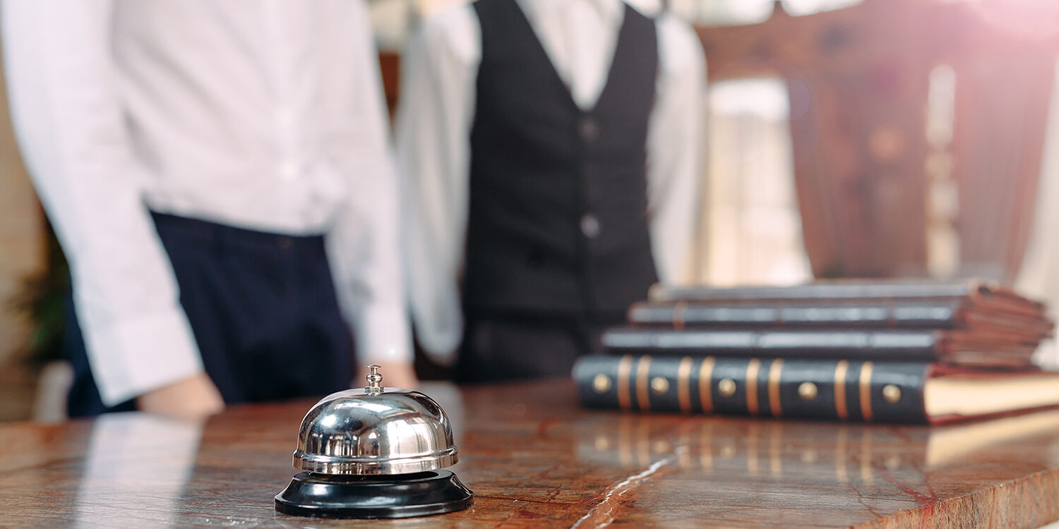 Diminuiscono i pernottamenti negli hotel nel 2020. Nella foto: la reception di un hotel con due receptionist in abiti eleganti. Il primo piano il campanello, tutto il resto è fuori fuoco
