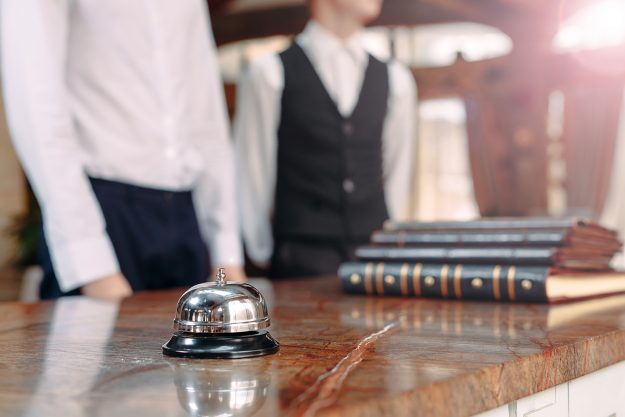 Diminuiscono i pernottamenti negli hotel nel 2020 Diminuiscono i pernottamenti negli hotel nel 2020. Nella foto: la reception di un hotel con due receptionist in abiti eleganti. Il primo piano il campanello, tutto il resto è fuori fuoco